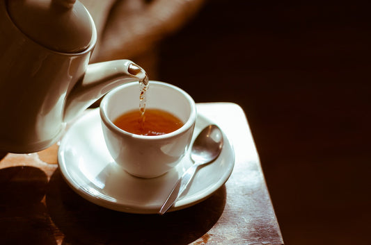 Teapot pouring hot tea into a ceramic cup on a saucer.